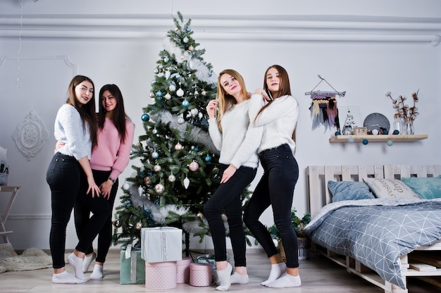 Cuatro lindas amigas se visten con suéteres calientes, pantalones negros contra el árbol con decoración navideña en la sala blanca.