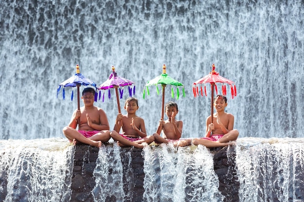 Cuatro muchachos balineses con sombrillas tradicionales indonesias meditan en una cascada.