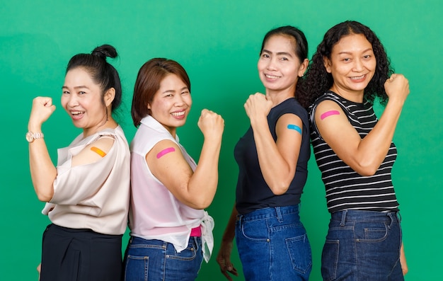 Cuatro mujeres asiáticas de mediana edad que muestran sus brazos con un parche de vendaje que muestra que se vacunaron contra el virus Covid 19 sobre fondo verde. Concepto de vacunación contra Covid 19.