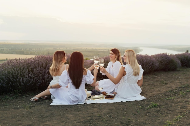 Cuatro mujeres vestidas de blanco están sentadas sobre una manta y brindando con copas de vino.