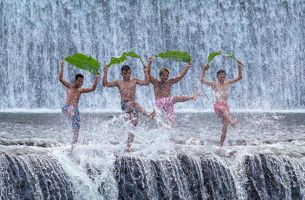 Cuatro niños balineses con grandes hojas de palma juegan con salpicaduras de agua cerca de una cascada