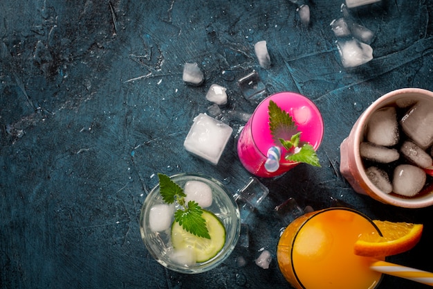 Foto cuatro tipos de bebidas refrescantes con hielo sobre un fondo azul oscuro y cubitos de hielo. concepto club nocturno, vida nocturna, fiesta, sed. naranja, menta y pepino, fresa, cola. vista plana, vista superior