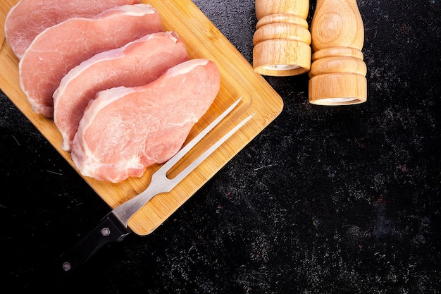 Cuatro trozos de carne de bistec crudo sobre una tabla de madera junto a dos saleros y pimenteros sobre un fondo de madera negra