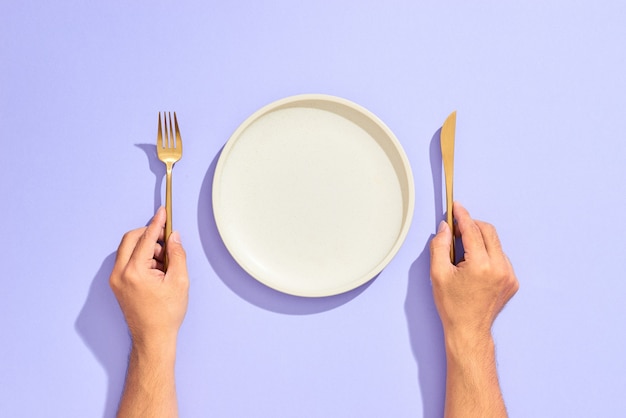 Cubierto de cena. Plato vacío blanco y mano sosteniendo tenedor y cuchillo de oro en lila