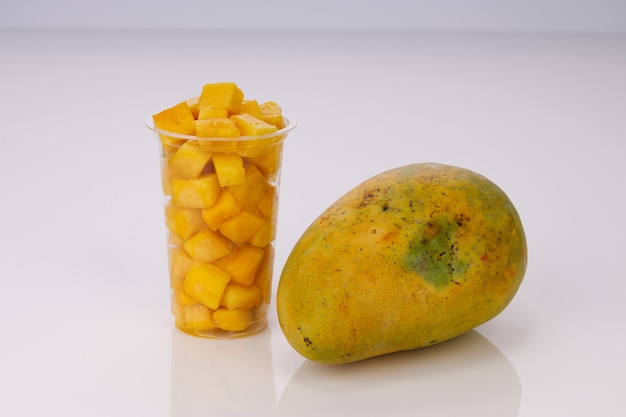 Cubitos de mango maduro o pieza cortada dispuestas en un vaso transparente con mango colocado cerca sobre fondo de color blanco.