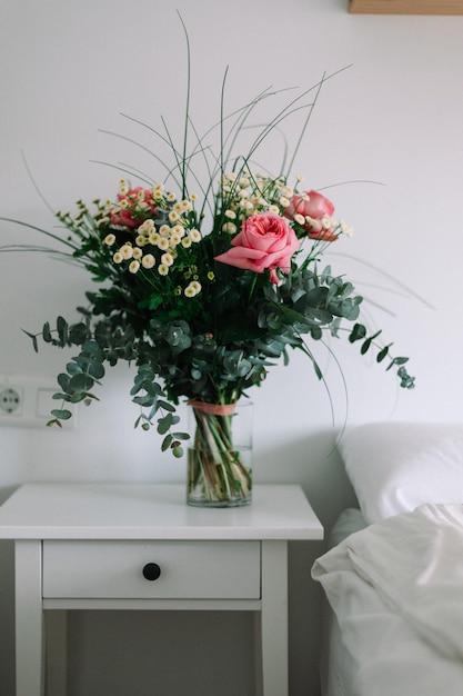 Cubo de flores silvestres sobre una base en la mesa de noche junto a la cama blanca. estilo nórdico.