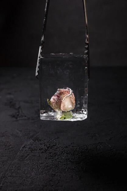 Un cubo de hielo transparente con una flor de rosa congelada en el interior en pinzas de hielo de camarero, fondo oscuro