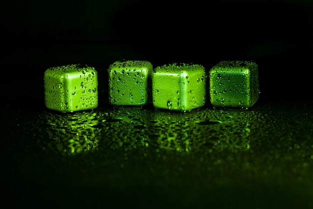 Cubos de aço inoxidável verdes simulando gelo para bebidas refrescantes em uma superfície preta com um reflexo.