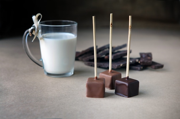 Cubos de chocolate no palito com copo de leite e cacau aromático e chocolate no fundo marrom. Cacau quente com leite em copo de vidro transparente, cubos de chocolate partidos de avelã