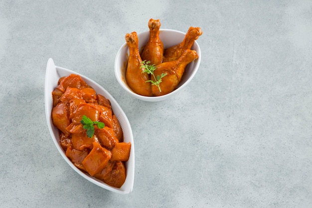 Cubos de frango marinado cru e pernas dispostas em uma louça de cerâmica branca guarnecida com coentro fresco e folha de hortelã que é colocada no plano de fundo texturizado branco cinza, foco seletivo.