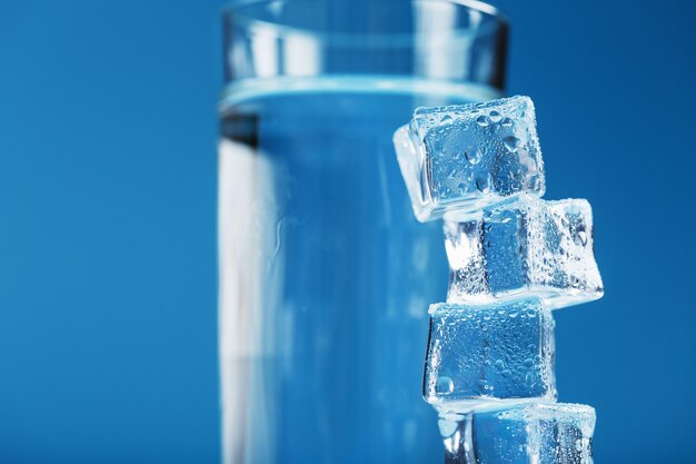 Cubos de gelo elevam-se com um copo de água fria e limpa sobre um fundo azul. Espaço livre