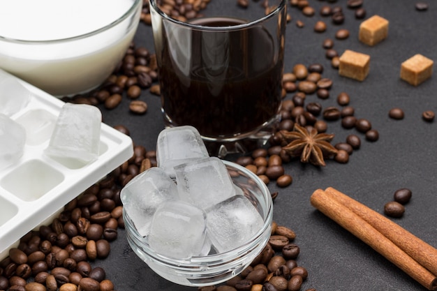 Cubos de gelo em pires, copo com café, xícara com leite. no fundo preto, grãos de café, pau de canela, anis estrelado.