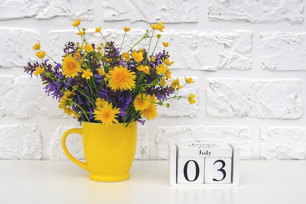 Cubos de madeira calendário 3 de julho e copo amarelo com flores coloridas brilhantes contra a parede de tijolos brancos.