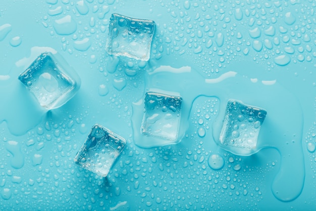 Cubos de hielo con gotas de agua esparcidos sobre un fondo azul, vista superior.