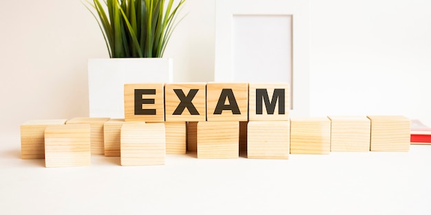 Cubos de madera con letras sobre una mesa blanca. La palabra es EXAMEN. Superficie blanca con marco de fotos y planta de interior