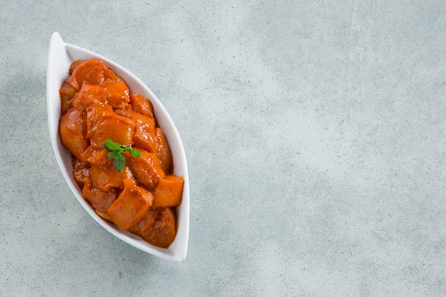 Cubos de pollo marinados crudos o pollo cortado en cubitos dispuestos en una vajilla de cerámica blanca adornada con hojas de cilantro frescas que se colocan sobre fondo de textura blanca gris.