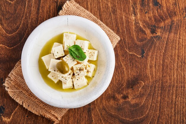 Cubos de queso feta con romero, aceitunas y salsa de aceite de oliva en un tazón blanco sobre la vieja mesa de madera marrón. Queso casero griego tradicional.