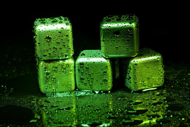 Cubos verdes de acero inoxidable que simulan hielo para enfriar bebidas sobre una superficie negra con un reflejo.