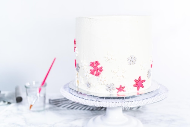 Cubriendo un pastel redondo alto con glaseado de crema de mantequilla italiana blanca y decorando con copos de nieve de fondant.