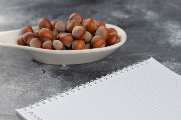 Cuchara blanca llena de avellanas saludables con cáscara con cuaderno abierto sobre mesa de piedra.