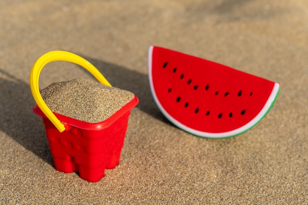 Cuchara de castillo de arena roja con juguete de sandía sobre arena.