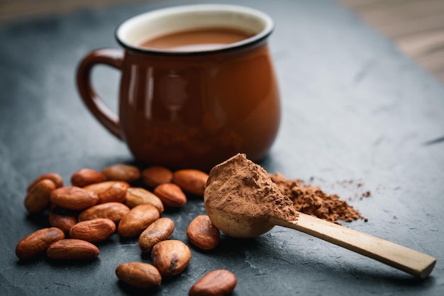 Cuchara de madera pequeña con cacao en polvo y granos de cacao con una taza marrón sobre un fondo