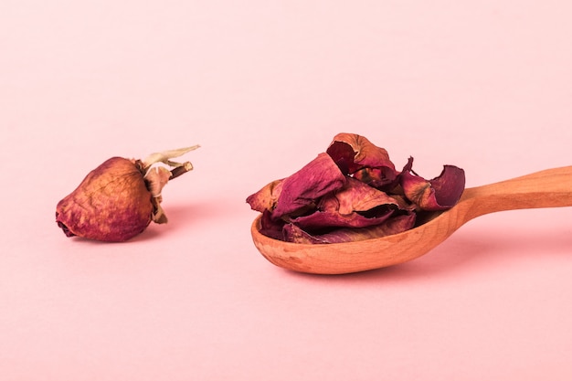 Una cuchara con pétalos secos y un capullo de rosa marchita sobre una superficie rosa.