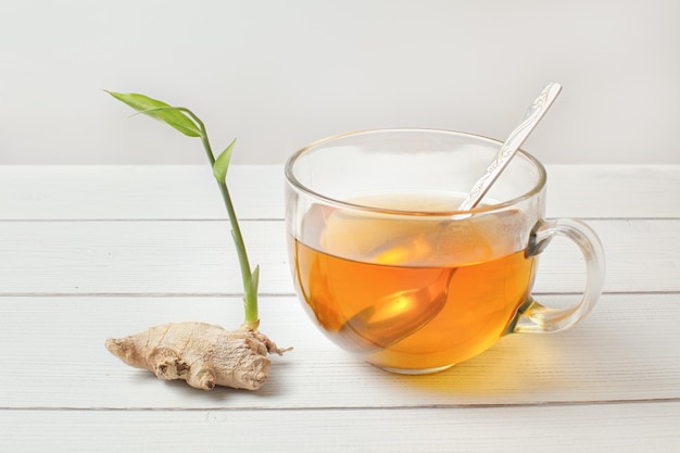 Cuchara en taza de vidrio de té ámbar caliente, raíz de jengibre seca con brote verde al lado.