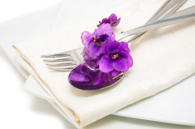 Una cuchara con un tenedor sobre una servilleta y un plato decorado con violetas.