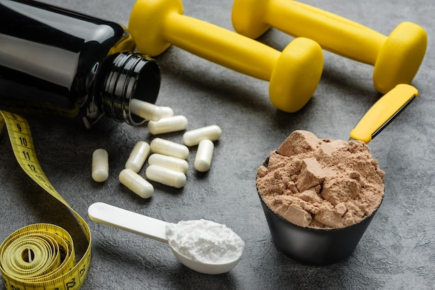 Cucharas medidoras con proteína de chocolate y creatina, cápsulas de aminoácidos, mancuernas y primer plano de cinta métrica. El concepto de suplementos deportivos.