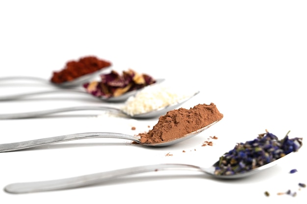 Cucharas de metal con diferentes ingredientes de especias de té aislados en blanco Copos de coco de cacao pétalos de rosa de lavanda