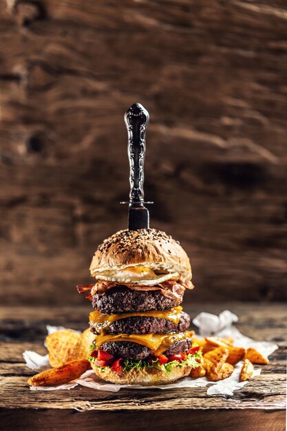 Cuchillo apuñalado en una hamburguesa triple con queso con empanada de ternera, queso derretido, huevo y pimientos en un ambiente rústico de madera.