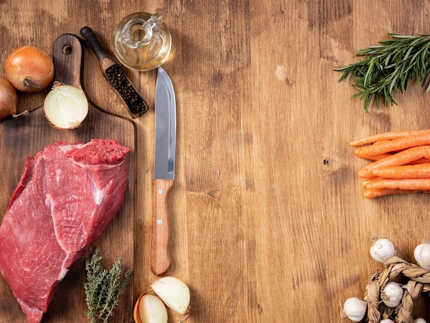 Cuchillo de chef junto a un gran trozo de carne roja y verduras frescas. Tabla de cortar de madera. Ajo en conserva.