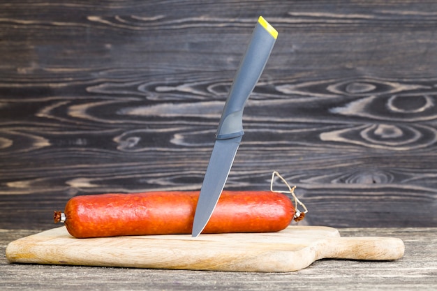 Cuchillo negro afilado y todo un palo de salchicha sobre una tabla de cortar de madera, comida en casa