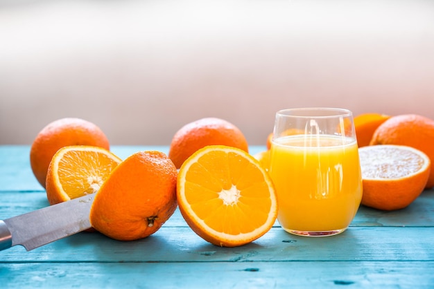 Cuchillo partiendo una naranja por la mitad, vaso de jugo natural de varias naranjas orgánicas, algunas enteras, algunas exprimidas, para la dieta, sobre una mesa de madera azul, Desayuno saludable