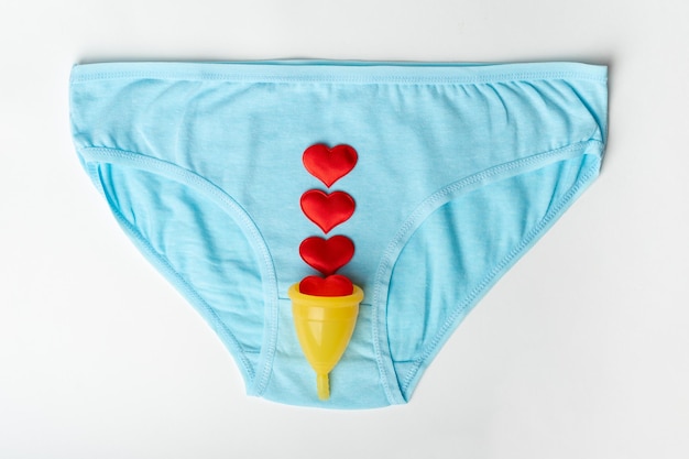 Cueca de calcinha azul feminina e um copo menstrual amarelo com corações vermelhos na forma de gotas menstruais em um fundo branco Conceito de saúde da mulher Conceito ecológico Conceito de desperdício zero