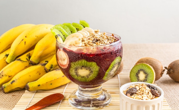 Cuenco de acai con copos de kiwi, plátano y avena sobre una mesa con decoración tropical.