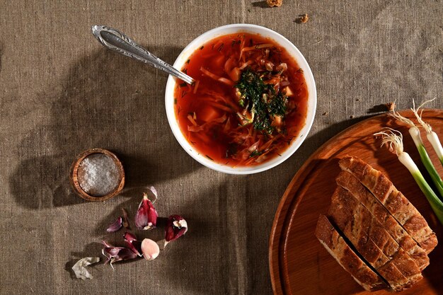 Cuenco de cerámica blanca con borscht, cebollas verdes y pan de molde sobre tabla de cortar de madera, ajo pelado y recipiente de sal sobre mantel de lino. Fondo plano de alimentos laicos. Cocina tradicional ucraniana