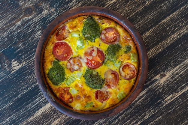 Cuenco de cerámica con frittata de verduras, comida vegetariana sencilla. Frittata con huevo, tomate, pimiento, cebolla, brócoli y queso en la mesa de madera, de cerca. Tortilla de huevo italiana