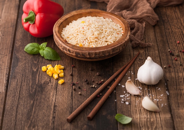 Cuenco de madera con arroz basmati de grano largo hervido con verduras sobre fondo de mesa de madera con palitos y pimentón rojo con maíz, ajo y albahaca.