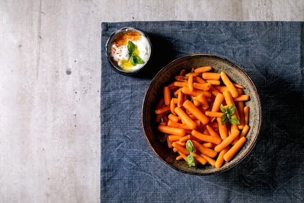 Cuenco de zanahoria baby entera al horno con salsa de yogur sobre un mantel azul sobre fondo gris. Cena vegana. Endecha plana, espacio de copia