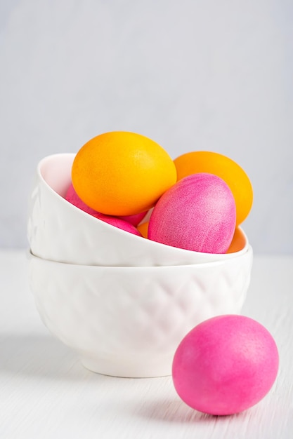 Cuencos de cerámica con coloridos huevos de pascua hervidos de color rosa y amarillo servidos sobre fondo de madera blanca