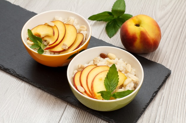 Cuencos de porcelana con gachas de sorgo, melocotón, anacardos, almendra y hojas de menta sobre tablero de piedra. Ensalada vegana de sorgo sin gluten con frutas frescas. Vista superior.