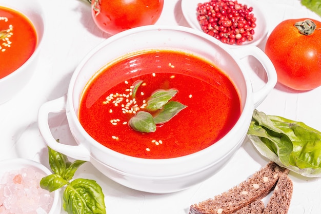 Cuencos con sopa de tomate casera. Verduras maduras, hojas frescas de albahaca, palitos de pan, especias aromáticas. Luz dura de moda, sombra oscura. Fondo de masilla blanca, endecha plana