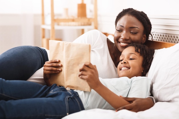 Cuento antes de dormir. Feliz mamá negra y su hijo leyendo un libro juntos, acostados en la cama en casa, espacio de copia