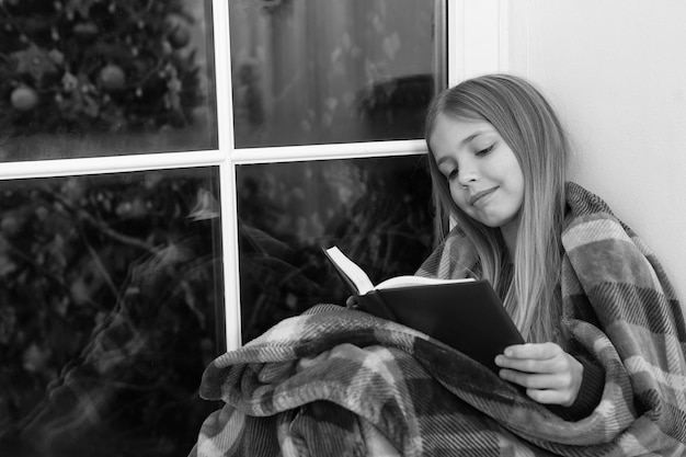 Cuento de hadas para felices fiestas Pequeño lector envuelto en tela escocesa sentado en el alféizar de la ventana Niña pequeña disfruta leyendo la historia de Navidad Niño pequeño lee el libro en la víspera de Navidad Libro para niños Espíritu navideño mágico