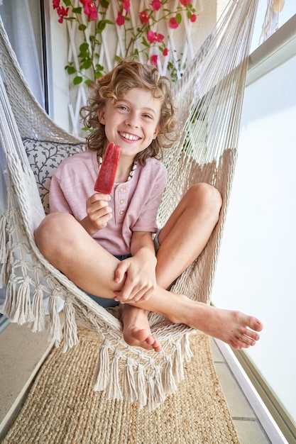 Cuerpo completo de alegre niño descalzo sonriendo y mirando la cámara mientras descansa en una hamaca de punto con las piernas cruzadas y comiendo delicioso helado en el balcón de casa