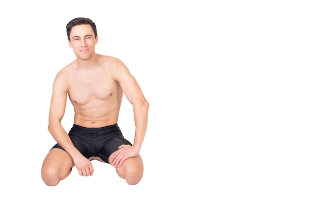 Cuerpo completo de deportista de contenido con el torso desnudo mirando a la cámara mientras se sienta aislado en fondo blanco en estudio de luz