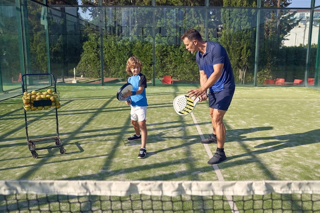 Cuerpo completo de entrenador masculino enseñando al niño a golpear con raqueta durante el entrenamiento de pádel en el patio de juegos con red y pelotas