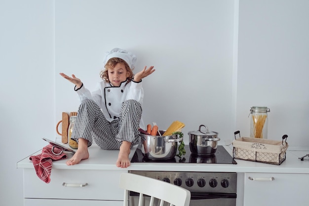 Cuerpo completo de un lindo niño preadolescente descalzo con uniforme de chef y gorra sentado en el mostrador de la cocina cerca de una estufa moderna con una cacerola desconcertado